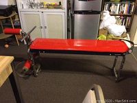 Red padded workout bench with metal frame and leg press attachment in carpeted room interior