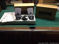 Two Harman Kardon black computer speakers with power adapter and original packaging, displayed on a green felt surface, with boxes and manuals.