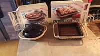 Two Pyrex baking dishes with original boxes on a kitchen counter. One is a 1.5 quart covered casserole with a dark lid in a rattan basket. The other is an 8x20 cm square cake dish with a glass insert and a rattan basket.
