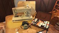 Portable Kenmore sewing machine with beige hard case and foot pedal on table, with container of thread and bobbins.