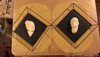 Pair of ceramic or stone face wall hangings shown together on table with woven placemats, displaying size and design.