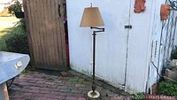 Vintage floor lamp with beige pleated lampshade, brass pole, and marble base shown outdoors beside a white shed and wooden fence.
