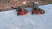 Two red Homelite chain saws on outdoor carpet showing wear and rust on blades