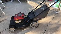 Side view of Craftsman lawnmower showing red engine cover, black metal deck, rear grass collection bag, and wheels on concrete surface.