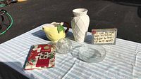 Overview photo of all decorative items displayed on a surface including vases, pitcher, bowl, paperweight, tablecloth in packaging, sign, and booklet