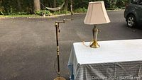 Photo of brass tone floor lamp and brass tone table lamp on a table with white tablecloth outdoors.