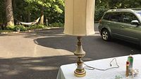 Photo of metal table lamp on white table with cream-colored fabric shade, taken outdoors showing full lamp, shade is tall and cylindrical with visible wear.