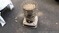 Front-angle view of the Sears kerosene heater, showing metal protective cage and base with control knobs, placed outdoors on pavement with scattered leaves and debris.