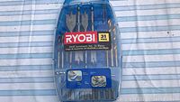 Front view of Ryobi 31-piece drill accessory set in closed transparent and blue plastic case showing drill bits and spade bits inside.
