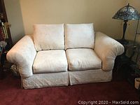 Full front view of the loveseat showing off white fabric with subtle palm leaf pattern, two seat cushions, two back cushions, rolled arms, and fabric skirt.