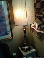 Brass table lamp with cream lampshade shown lit beside framed artwork and textile.