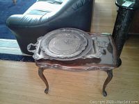 Wooden side table with glass top and two tarnished metal trays placed on top, next to black leather couch and hardwood floor visible