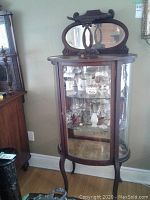 Front full view of antique curio cabinet showing the curved glass front panel and wooden frame with the ornate top and mirror.