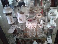 Overview of seven-piece Hungarian porcelain lot including lamp, watering can, trinket box, vases, and drinking glasses displayed in glass cabinet