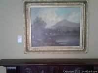 Framed landscape painting shown hanging on wall above carved wood furniture