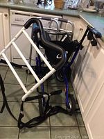 Photo of two walkers and one bed assist rail on the kitchen floor, showing the seat and storage pouch attached to blue walker and the white bed assist rail in front.