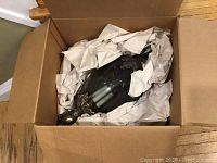 Light fixture partially wrapped in packing paper inside box, showing dark glass and metal parts.