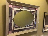 Full view of stained glass mirror with wood frame against beige wall showing oval center mirror and surrounding decorative stained glass panels in purple, pink, and white.