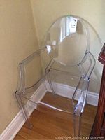 Clear Lucite chair seen from front corner angle, showing curved backrest and armrests, standing on wood floor.