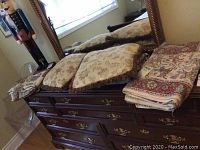 Multiple floral throw cushions and folded linens on wooden dresser beneath a mirror.