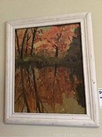 Full view of the oil painting in a white frame showing autumn trees with reflections.