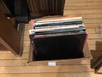 Vinyl records stacked in a cardboard box showing multiple records with various colored sleeves.