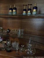 Photo shows decorative blue and gold glasses with twisted gold stems on upper shelf and various glassware on lower shelf including small bowls and glasses.