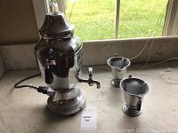 Electric metal coffee carafe with attached power cord on counter with two metal cups.