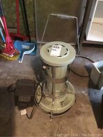 Aladdin cylindrical kerosene heater on floor, showing top handle and protective metal cage around burner