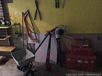 Photo of assorted garden tools hanging on pegboard including seed spreader cart, red Toro electric hedge trimmer, white Craftsman leaf blower, and red metal tool boxes.