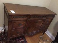 Front and angled view of antique wooden chest showing two drawers on the left side, metal ring pulls, decorative carvings, dark wooden veneer with some chips, standing on tapered legs.