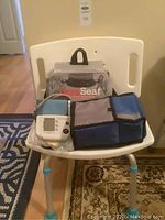 White Lava shower seat with curved backrest. Life Source blood pressure monitor, cuff, and soft carrying bag placed on seat. Brown floor and patterned rugs visible.