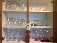 Upper and middle shelves of cabinet showing an assortment of tall clear glasses, stemmed wine glasses, small colored glass votives, and some pink glass items on a lower shelf.