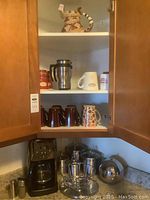 Upper and lower cabinet shelves showing mugs, ceramic pitcher, teapot shaped like an animal, and kitchen items including coffee maker and canisters.