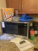 Photo showing Panasonic microwave with popcorn buckets on top, blue plastic party plates and cutlery, colorful plastic cups, Cuisinart coffee maker to the right, clear travel mug, and white plastic plates in front on countertop.
