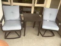 Full set showing two metal outdoor chairs with light gray cushions and one matching metal side table between them on a concrete surface.