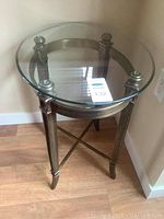 Round side table with clear glass top and heavy metal legs on wooden floor.