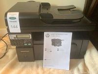 Full view of HP LaserJet Professional M1210 printer showing the top automatic document feeder and control panel with a MaxSold lot tag and printed user manual in front.