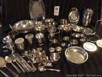 Full view of mixed stainless steel and silver plate serveware set including bowls, serving trays, condiment holders, and cutlery on black cloth.
