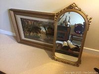 Framed landscape print and ornate framed beveled mirror leaning against a wall on carpet.