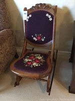 Front view of the folding rocking chair showing carved wood and needlepoint floral upholstery on backrest and seat.