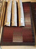 Four rolled carpets placed on top of one unrolled carpet showing burgundy and brown gradient plaid pattern.