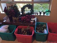 Four storage bins filled with assorted Christmas decorations including artificial wreaths and floral picks on table and floor near window