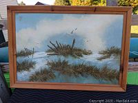 Framed oil painting of a beachscape with sand dunes, grasses, broken wooden fences, a cloudy sky, and flying seagulls.