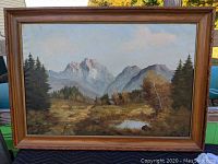 Full view of the framed oil painting depicting a snow-capped mountain landscape with trees and a small pond in the foreground under a cloudy blue sky.