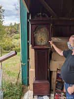Full view of the grandfather clock standing tall next to a man, showing the entire wooden case and clock face, missing one leg on the case base.