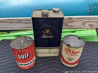 Photo showing three vintage metal advertising cans from Gulf, Standard, and Texaco, placed on a blanket outside with a wooden fence and blue vehicle in the background.