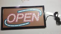 Front view of electric lighted 'OPEN' sign showing pink and blue neon-style tubing inside black frame with power cord attached.