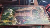 Full view of large framed print depicting a rural covered bridge scene with child, dogs and church steeple under bright sky.