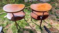Pair of kidney-shaped leather-top wooden end tables shown outdoors on grass, highlighting overall condition and shape.